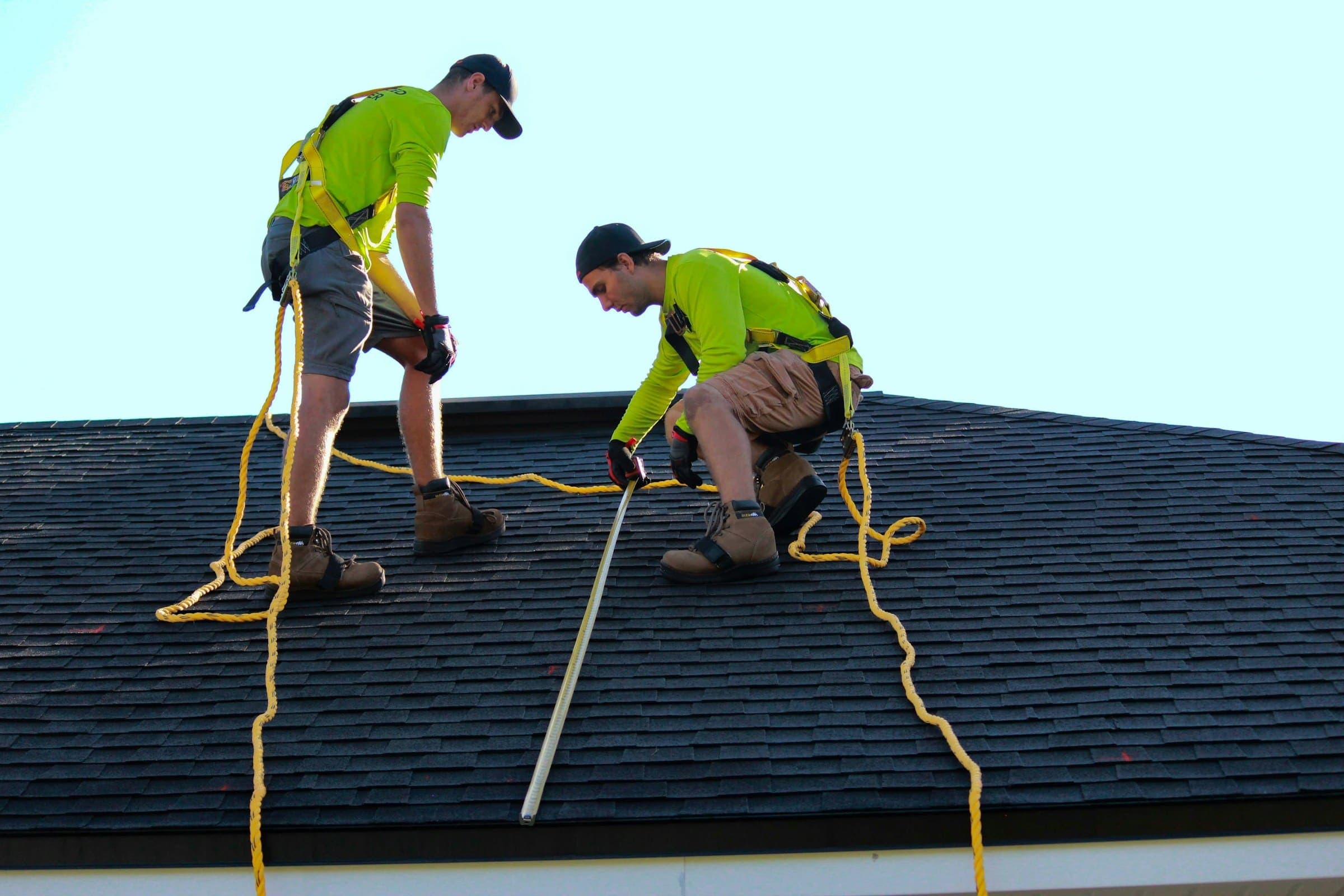 Two roofers in safety harnesses tied off while working on a shingle roof